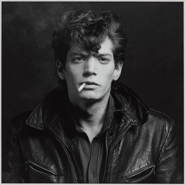 Robert Mapplethorpe, Self Portrait, 1980. Robert with a cigarette in his mouth and a serious face while wearing a polo under a leather jacket