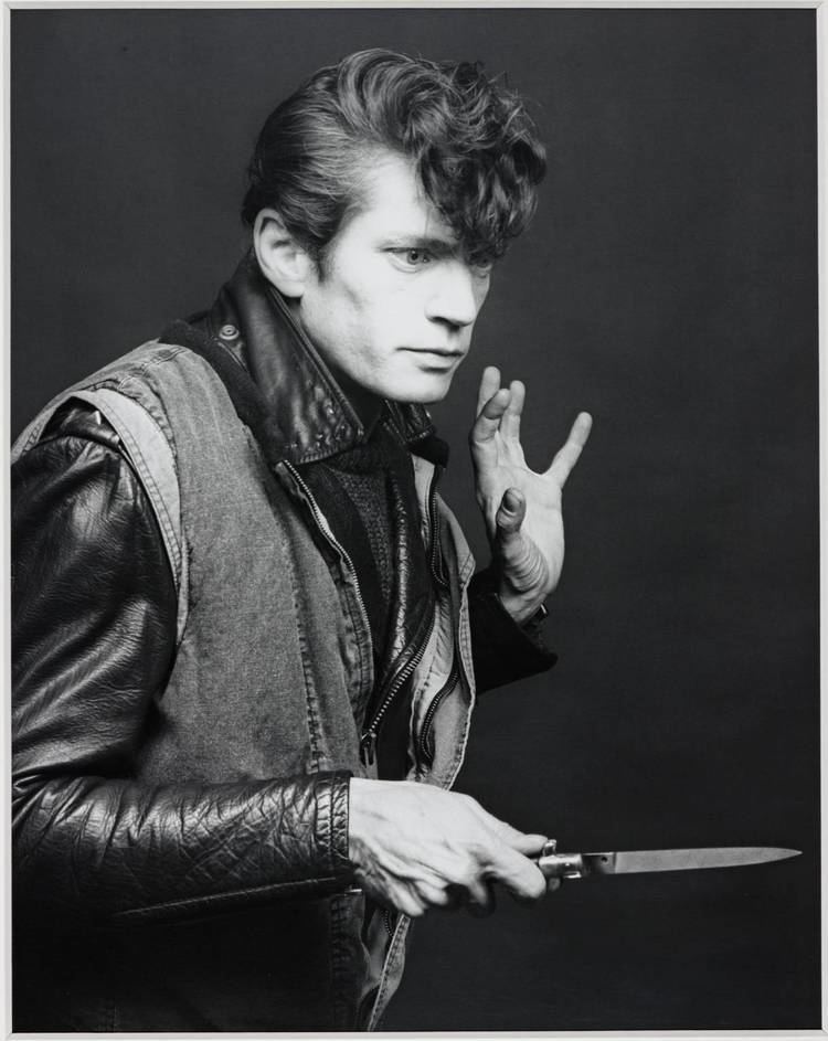 Self-Portrait, Robert Mapplethorpe, 1983. Robert holding a knife, with a surprised look, while wearing a shirt under a leather jacket and a sleeveless jacket