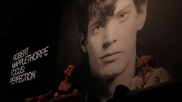 A woman and a man smiling and looking at each other while sitting in front of a big monitor where Robert Mapplethorpe's serious face is showing and a caption on the left side of the monitor says, "Robert Mapplethorpe focus perfection". The woman is wearing a black blouse while the man is wearing a  white long sleeve with eyeglasses attached to his shirt