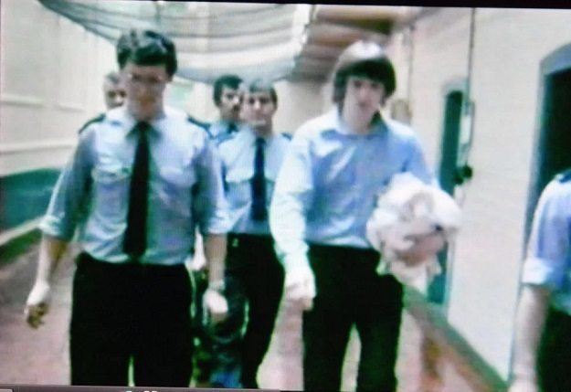 Robert John Maudsley wearing blue long sleeves escorted by five guards in their blue uniform.