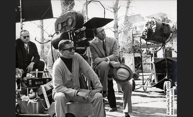 Robert Mulligan smiling and looking at something and behind him on the left is a man and on the right is Gregory Peck holding a hat with cameras on the background on the set of the 1962 American drama film, To Kill a Mockingbird. Robert is wearing sunglasses, a ring, pants, and a long sleeve shirt under a cardigan. The man on the left is wearing sunglasses, pants, a wristwatch, and a shirt under a long sleeve.  Gregory is wearing eyeglasses, pants, shoes, and a long sleeve under a necktie and coat.