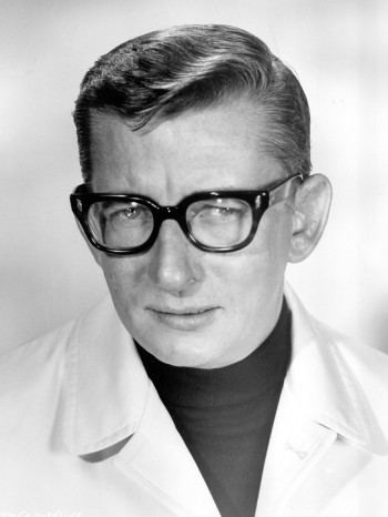 Robert Mulligan with a tight-lipped smile while wearing eyeglasses and a turtle-neck shirt under a coat