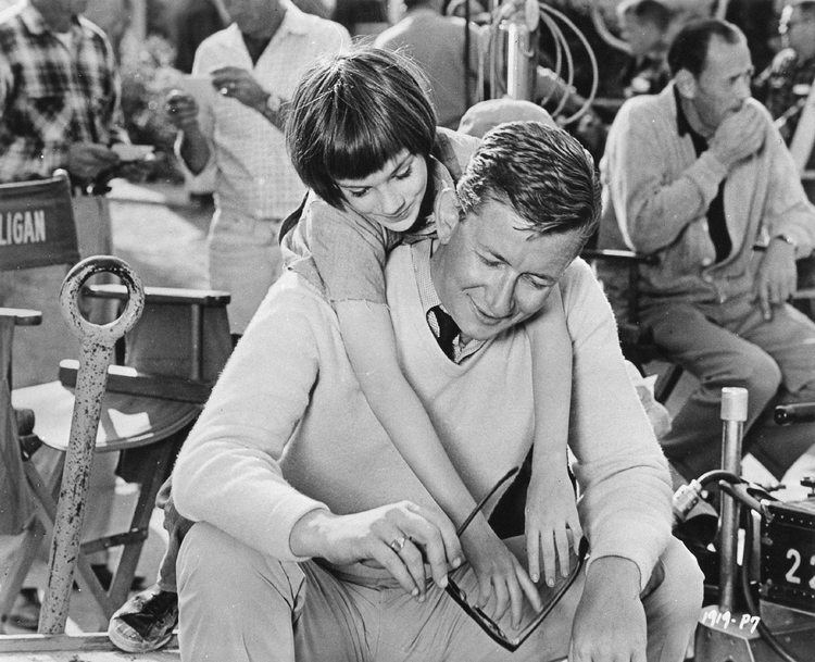 Mary Badham smiling and hugging Robert Mulligan while reaching the eyeglasses with people in the background on the set of the 1962 American drama film, To Kill a Mockingbird. Mary is wearing a blouse and Robert is wearing pants and a long sleeve under a sweater