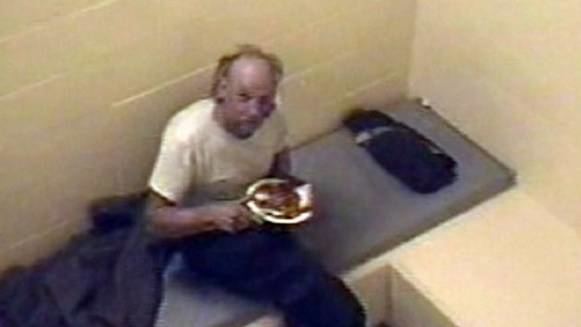 Robert Pickton holding a plate while sitting on the bed and wearing a white t-shirt and black pants