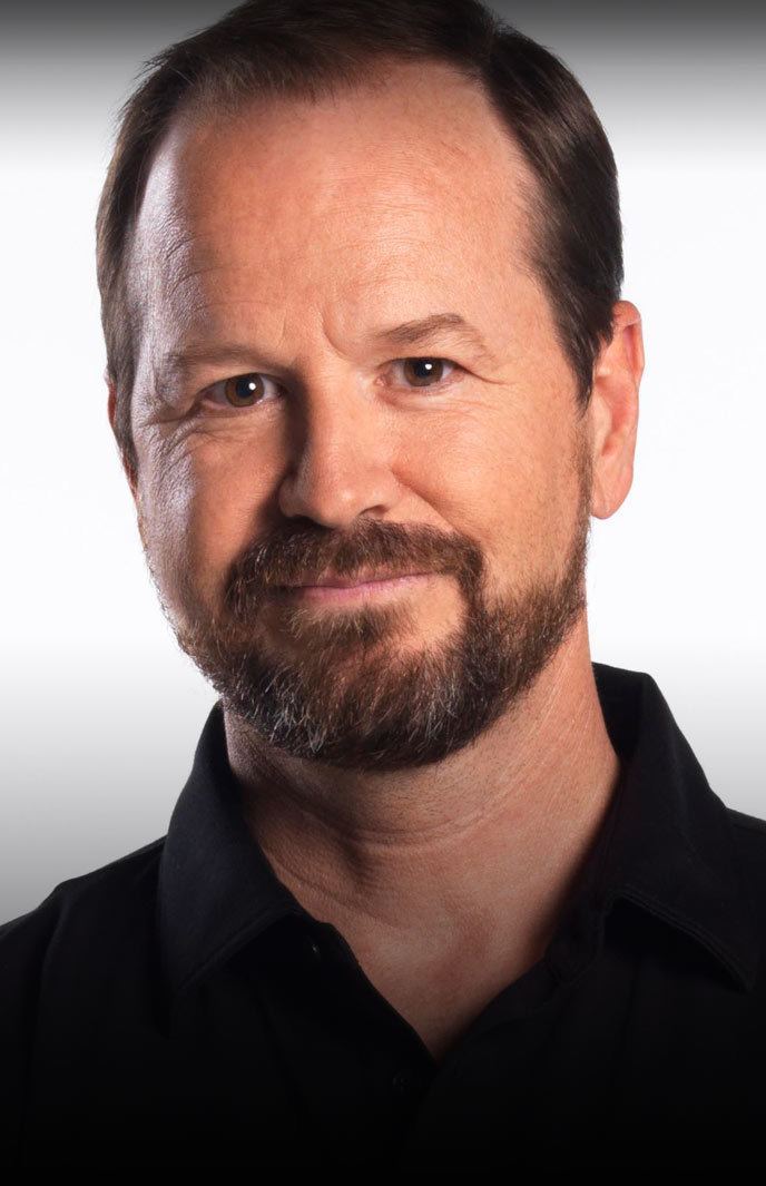 Robert Wahlberg with a tight-lipped smile, mustache, and beard while wearing a black polo shirt