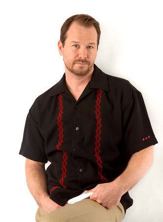 Robert Wahlberg with a tight-lipped smile, mustache, and beard while wearing a black and red polo and khaki pants
