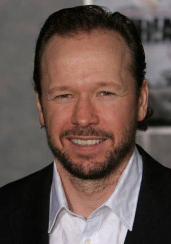 Donnie Wahlberg smiling with mustache and beard while wearing a black coat and white long sleeves