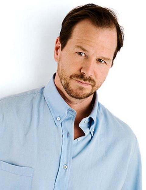 Robert Wahlberg with a tight-lipped smile, mustache, and beard while wearing a light blue long sleeves