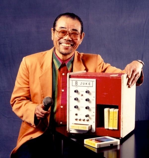 Daisuke Inoue smiling while holding a microphone and the Juke 8 karaoke machine on the table. Daisuke with a mustache and beard is wearing eyeglasses, a wristwatch, black pants, and a colorful long sleeve under an orange coat