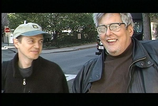 Rockets Redglare smiling and wearing a black leather jacket, eyeglasses, and brown shirt while Steve Buscemi wearing a black jacket