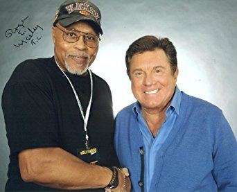 Roger E. Mosley smiling while wearing a black t-shirt, black cap, bracelet and necklace while Larry Manetti wearing blue long sleeves