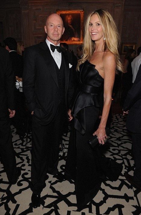 Roger Jenkins (left) is smiling, bald, standing, right hand inside his pocket, he is wearing white long sleeves, a black bow tie under a black suit with a pocket on left with a white handkerchief. Elle Macpherson is smiling, has long blonde wavy hair, and her left hand holding a black purse, she is wearing silver earrings black off-shoulder sexy dress. Behind them is a painting, surrounded by people.