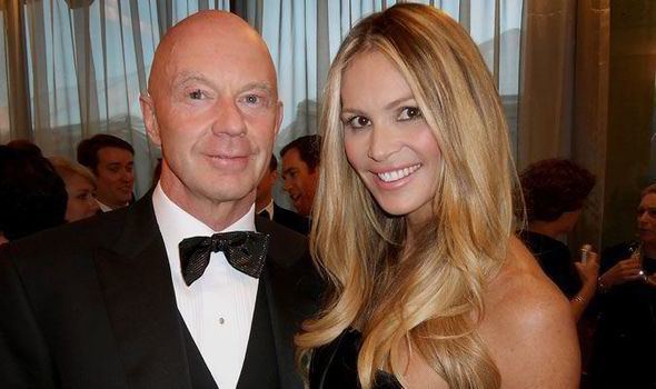 Roger Jenkins (left) is smiling, bald, and wears white long sleeves, a black bow tie under a black suit. Elle Macpherson (right) is smiling, and has blonde hair, wears a black sexy top. Behind them are people talking.