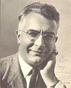Roger Peyrefitte with a tight-lipped smile while hand on his chin and wearing a coat, long sleeves, necktie, eyeglasses, and ring