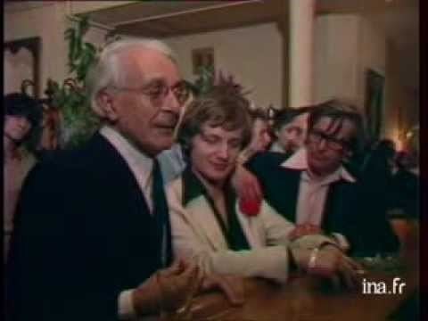 Roger Peyrefitte talking to someone with the people beside him while wearing a black coat, white long sleeves, necktie, and eyeglasses