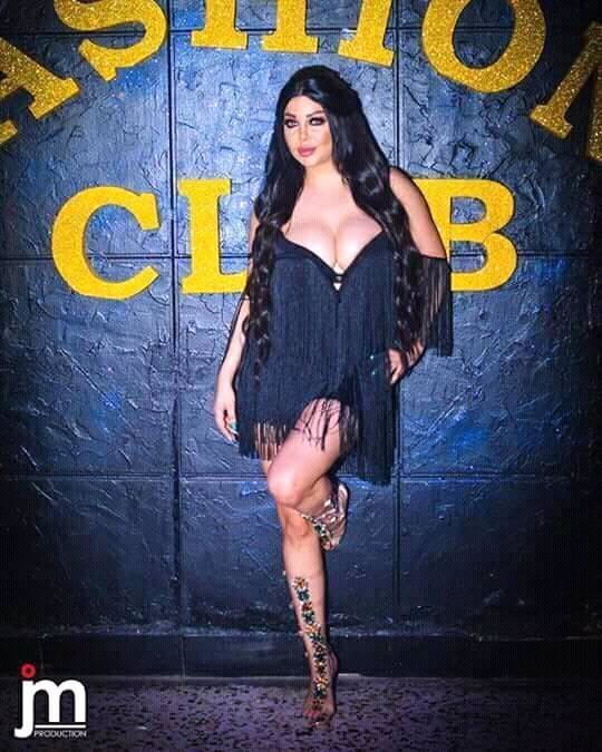 Rola Yamout posing in a wall with very long black hair and weaing a seductive black dress.