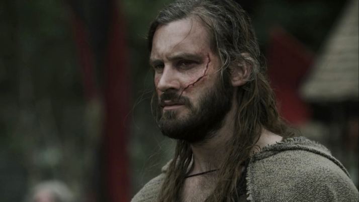 Clive Standen as Rollo looking serious in a scene from the 2013 film "Vikings" with a long hair, mustache, beard, and a wound on his face and wearing a gray cloak