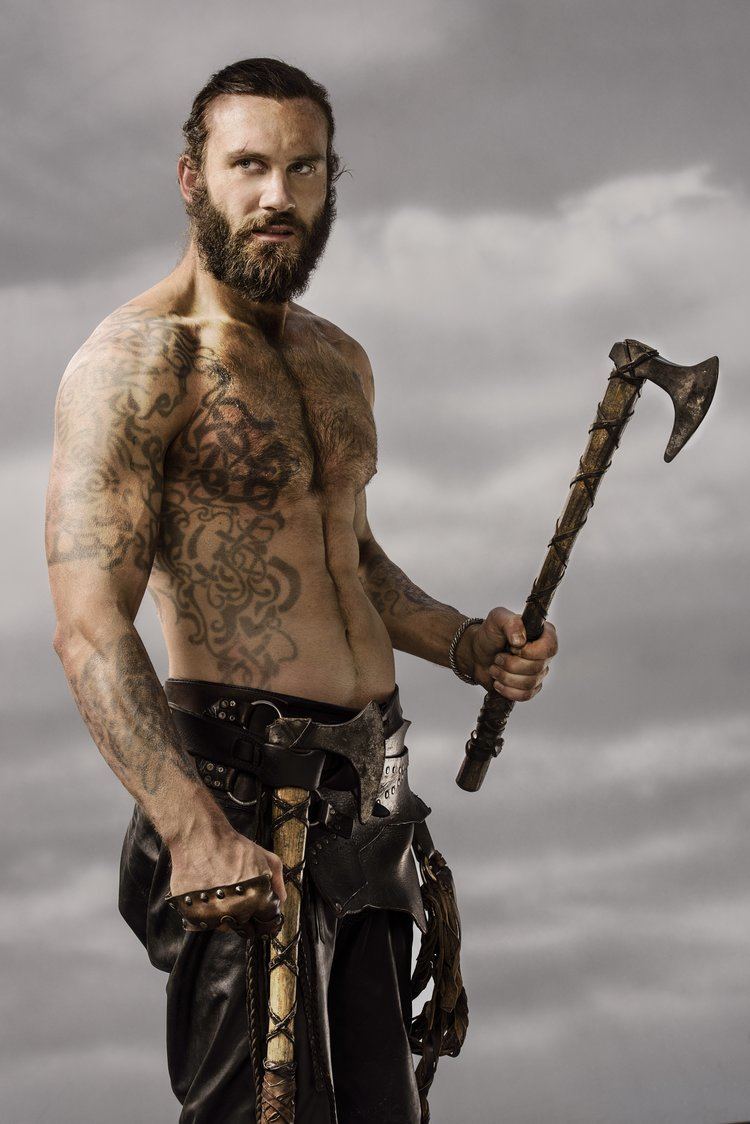 Clive Standen as Rollo looking serious in a scene from the 2013 film "Vikings" with short hair, beard, and mustache while holding an ax, topless and wearing black pants with another ax on the side of his belt and tattoos on his body