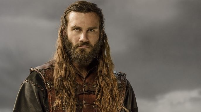 Clive Standen as Rollo looking serious in a scene from the 2013 film "Vikings" with long hair, mustache, and beard and wearing a warrior costume