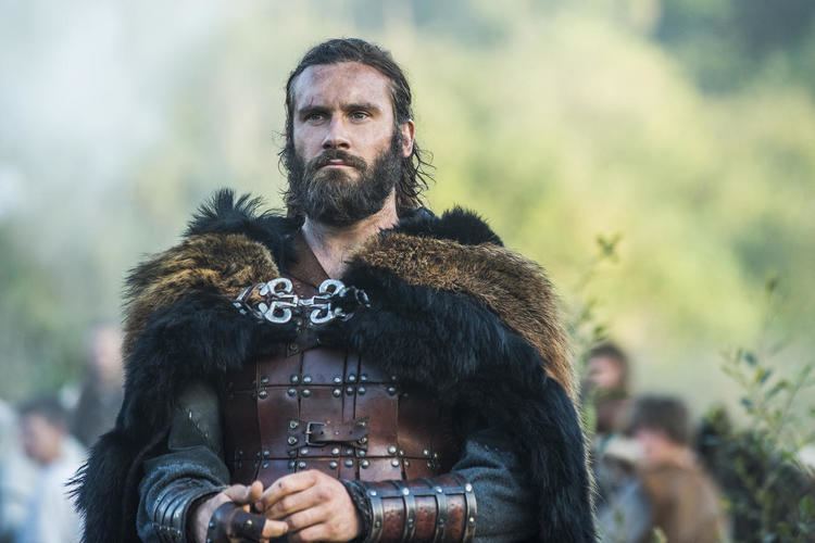 Clive Standen as Rollo looking serious in a scene from the 2013 film "Vikings" with a long hair, mustache, beard, and wearing a warrior suit under a black and brown cloak