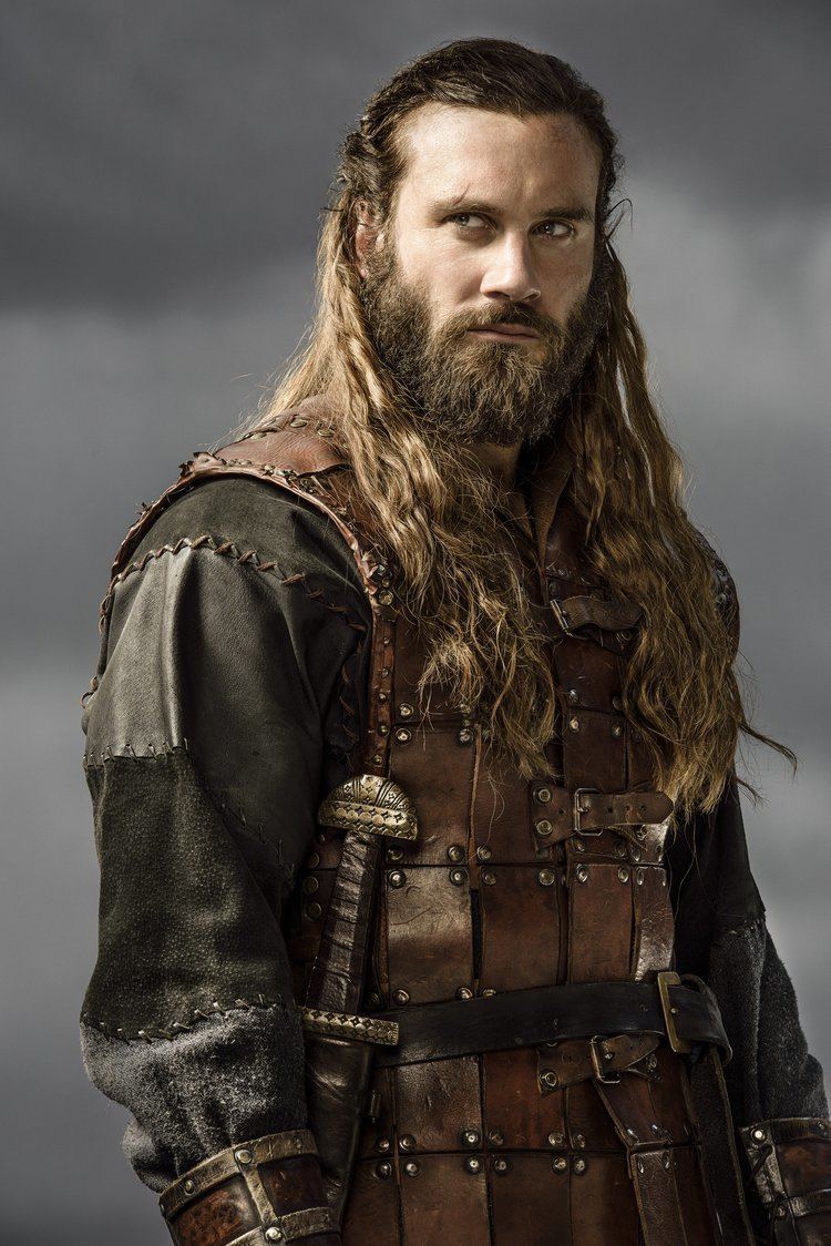 Clive Standen as Rollo looking serious in a scene from the 2013 film "Vikings" with a long hair, mustache, and beard and wearing a warrior suit with a sword attached on his belt