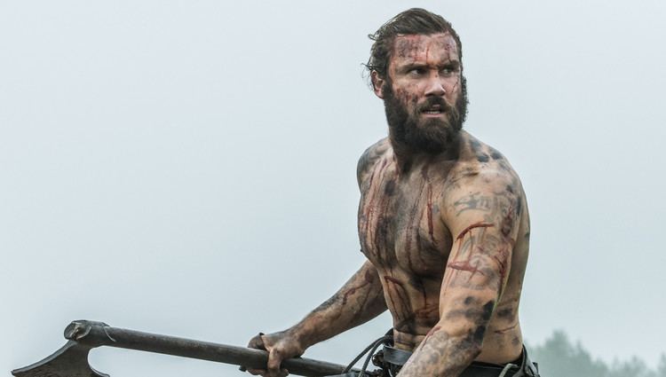 Clive Standen as Rollo looking serious in a fighting scene from the 2013 film "Vikings" with short hair and holding an ax, topless with tattoos and wounds on his body