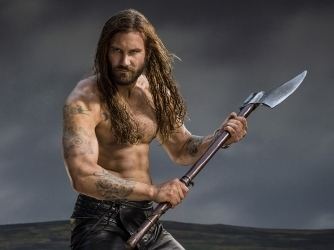 Clive Standen as Rollo looking serious and holding an ax in a scene from the 2013 film "Vikings" with long hair, mustache, beard and wearing black pants and topless