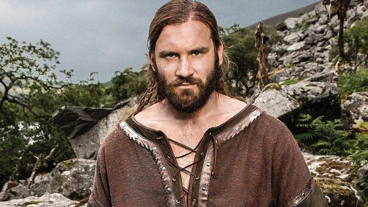Clive Standen as Rollo looking serious in a scene from the 2013 film "Vikings" with long hair, mustache, beard and wearing a brown clothing