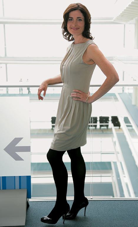 Romilly Weeks smiling in a side pose with left hand on the hips while wearing a beige sleeveless dress, black stocking, and black heels