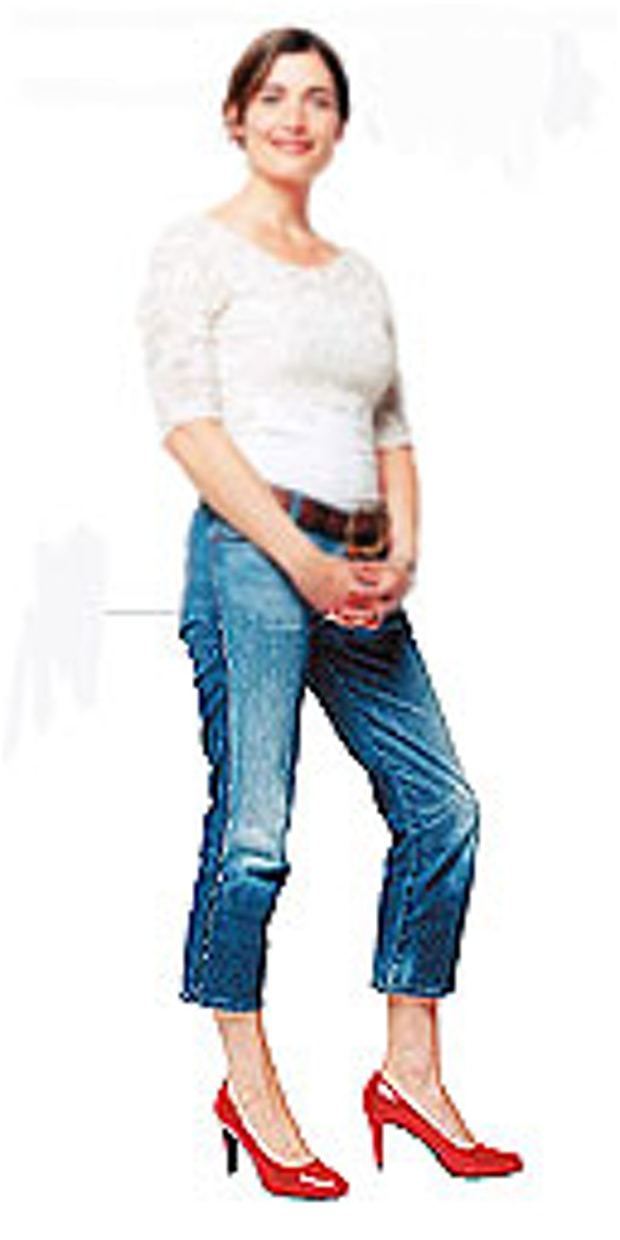 Romilly Weeks smiling in a standing pose and holding her own hands while wearing a white short sleeve blouse, high rise denim with a brown belt, and red heels