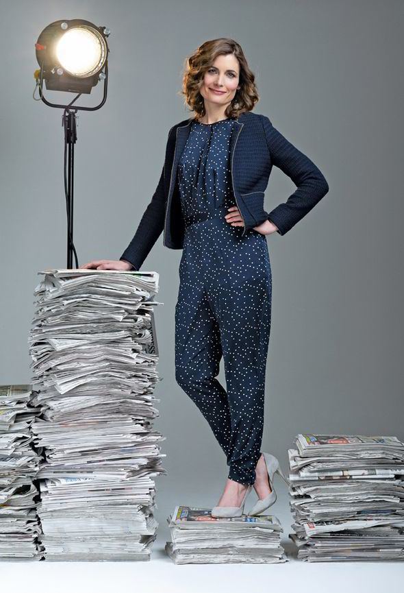 Romilly Weeks smiling and standing on a pile of newspapers with curly hair and left hand on the left hip while other hand touching another pile of newspapers with a lamp on top and wearing a polka dot jumpsuit under a blue coat and gray heels
