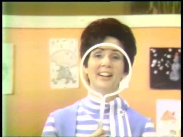 Miss Barbara in Romper Room wearing a striped white and blue shirt while holding a white circled object.