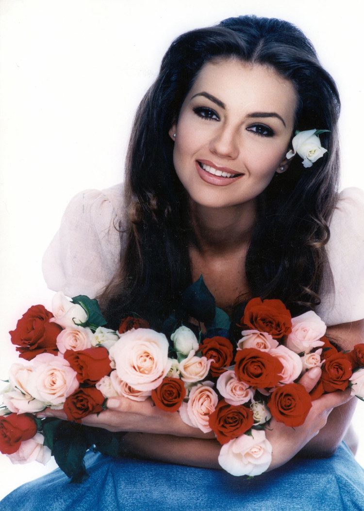 Thalía as Rosalinda, smiling while holding a bunch of flowers while wearing a white blouse and blue skirt
