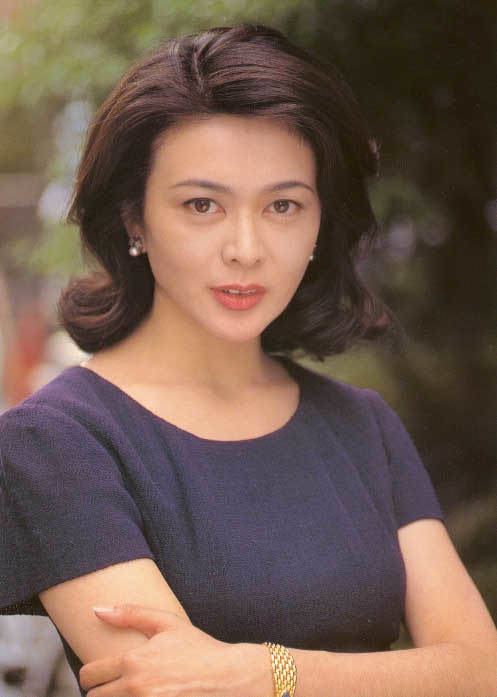 Rosamund Kwan smiling while wearing a dark blue blouse, earrings, and gold bracelet
