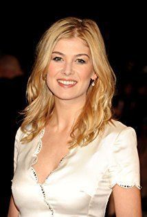 Rosamund Pike smiling with blonde wavy hair while wearing earrings and a white blouse with beads on the edges