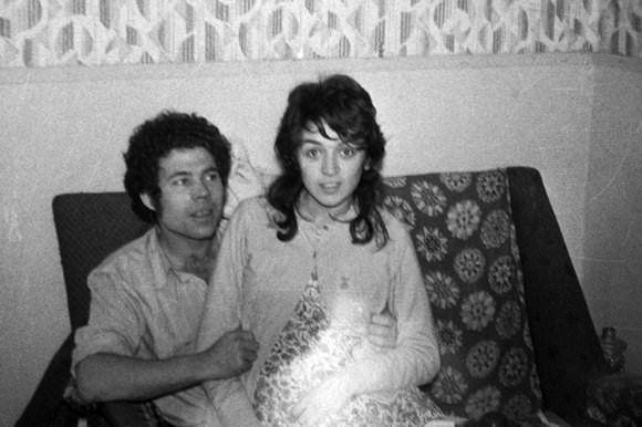Fred West is looking at Rosemary West sitting on his lap while smiling. Fred with curly hair and wearing a white folded long sleeves while Rosemary with wavy hair, wearing a white blazer over a floral dress.