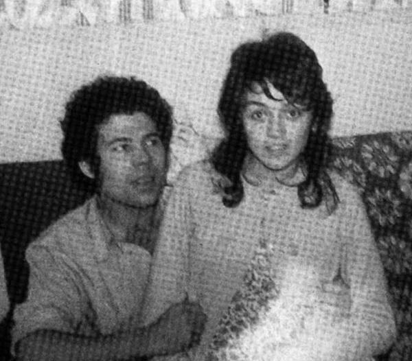 Fred West is looking at Rosemary West sitting on his lap while smiling. Fred with curly hair and wearing a white folded long sleeves while Rosemary with wavy hair, wearing a white blazer over a floral dress.