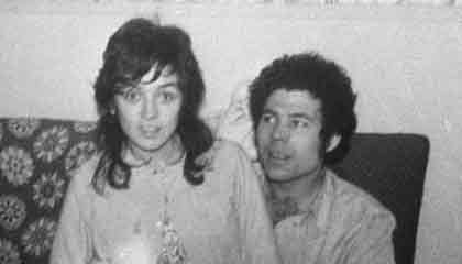 Fred West is looking at Rosemary West sitting on his lap while smiling. Fred with curly hair and wearing a white folded long sleeves while Rosemary with wavy hair, wearing a white blazer over a floral dress.