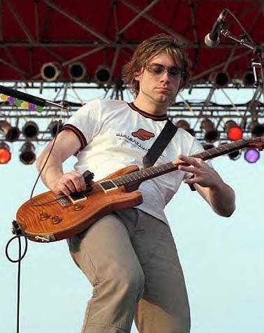 Ross Childress playing the guitar while wearing a white printed shirt, gray pants, and shades