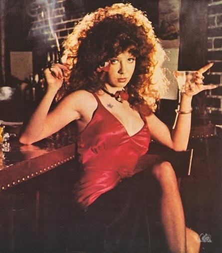 Ruby Starr with curly hair, wearing a necklace, bracelet, and a sexy red dress.