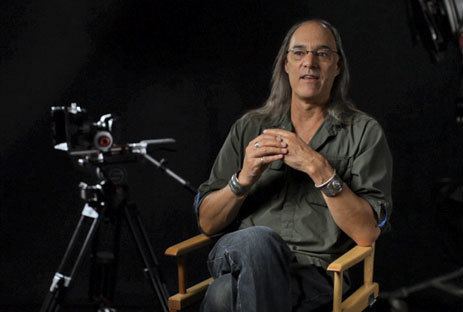 Russell Carpenter sitting on the chair while talking to someone and he is wearing eyeglasses, gray long sleeves, denim pants, and wristwatch