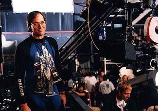 Russell Carpenter with a tight-lipped smile while on the set of Titanic and he is wearing eyeglasses and a black and blue rash guard