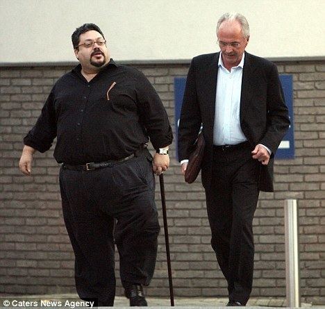 Russell King walking with a cane, wearing eyeglasses, a watch, a black long sleeve shirt, and black pants with Sven-Goran Eriksson carrying a handbag wearing a white shirt under a black coat, black pants, and eyeglasses