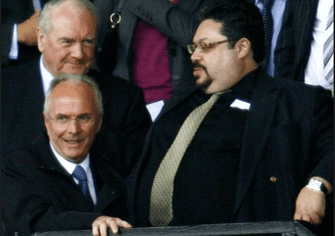 Russell King looking at Sven-Goran Eriksson with a beard and mustache and wearing a black shirt under a black coat, a necktie, and watch while Sven-Goran Eriksson talking and wearing a white shirt under a black coat, a blue necktie and eyeglasses