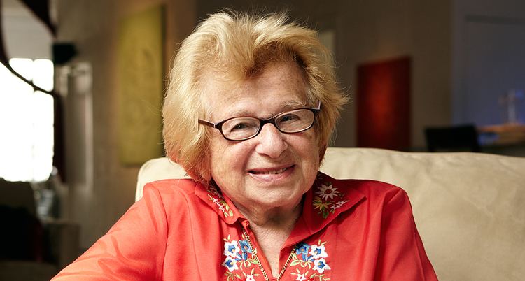 Ruth Westheimer in her beautiful smile and blonde hair while wearing eyeglasses and orange floral long sleeves