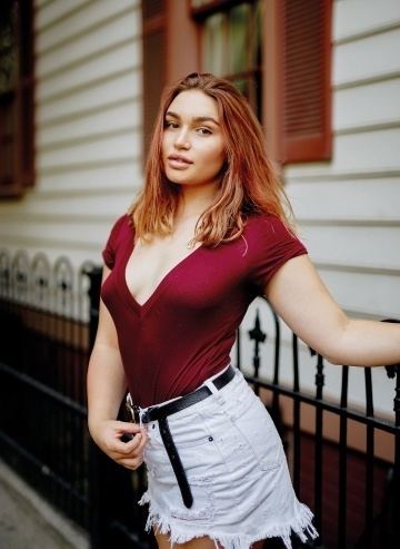 Rylee Martell smiling and holding into the fence while wearing a maroon blouse exposing her cleavage and white shorts with a black belt
