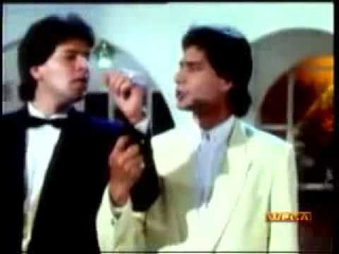 In a movie scene of SAATHI 1991, in a room with white walls and large rounded arc window, From left Aditiya Pancholi is serious standing, left hand up looking to his left, has black hair wearing a white polo with bow tie under a black coat, Moshin Khan is serious, speaking, mouth half open, standing, holding Aditiya’s arm, with his right arm has black hair wearing a white polo under a white cream coat.