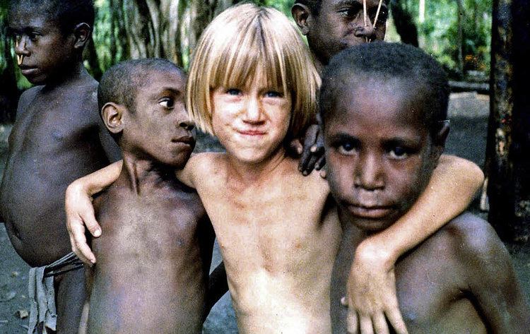 In a jungle, with native children, from left a a boy with a black skin, has black hair and a nose piercing, wearing a native cloth at the bottom, 2nd from left, a boy is serious, standing, looking at his left, has black hair naked, 3rd from left, Sabine Kuegler is smiling, standing with his hands behind over the shoulder of both kids on the side, has blond hair naked, 4th from left, behind a boys is serious, standing looking to the left has black hair has nose stick piercing naked, at the right, a boy is serious, standing, has black hair, top naked,