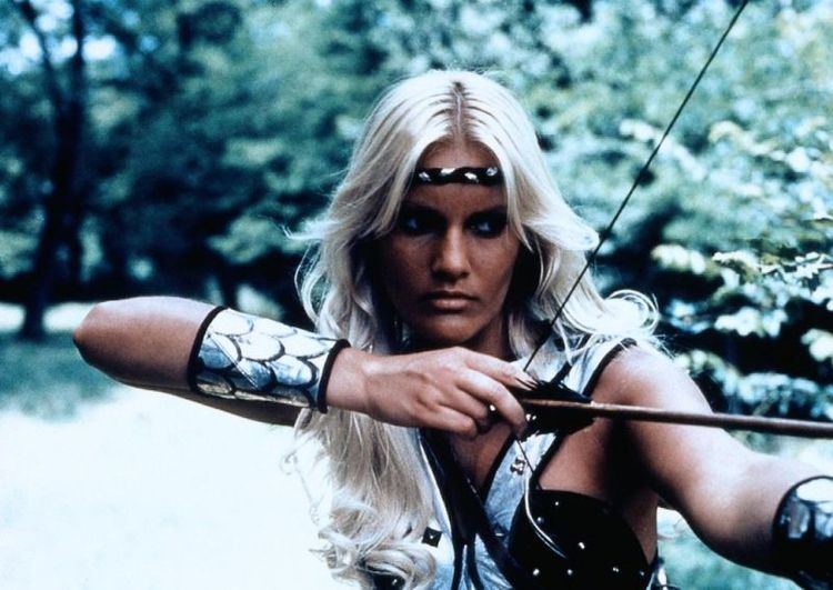 Sabrina Siani as Roon with a fierce look while holding an arrow and bow, with blonde hair, and wearing a white and black outfit in a movie scene from Ator, the Fighting Eagle, a 1982 Italian adventure-fantasy film.