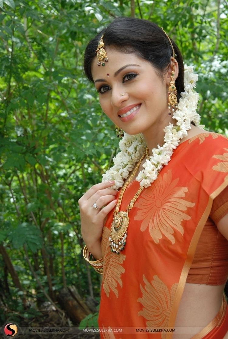 Sadha smiling with her hair arranged and wearing an orange colored choli top and sari as well as various jewelry and a garland of flowers.